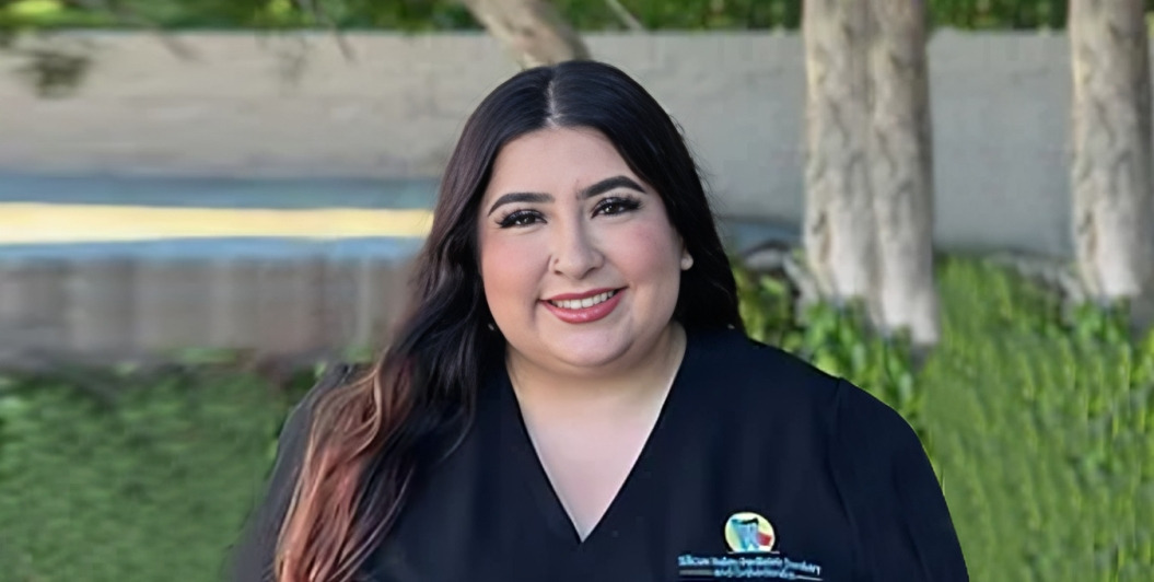 Woman in dark scrubs smiling outdoors with blurred dental office background.