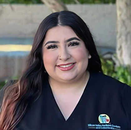 Woman in black medical scrubs smiling at camera, wearing dental clinic badge outdoors.