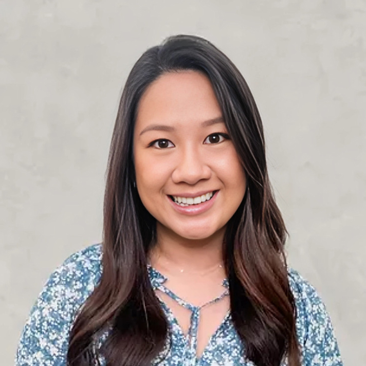 Professional headshot of smiling woman with long dark hair wearing blue patterned top against neutral background.
