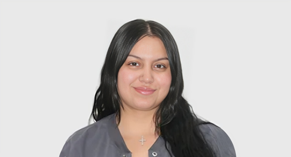 Professional headshot of woman with long dark hair wearing gray shirt against white background.
