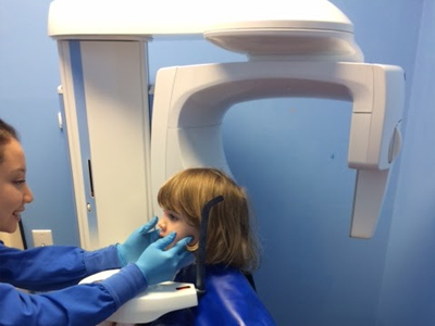 Pediatric dentist in blue scrubs positioning young patient's head for dental X-ray examination.