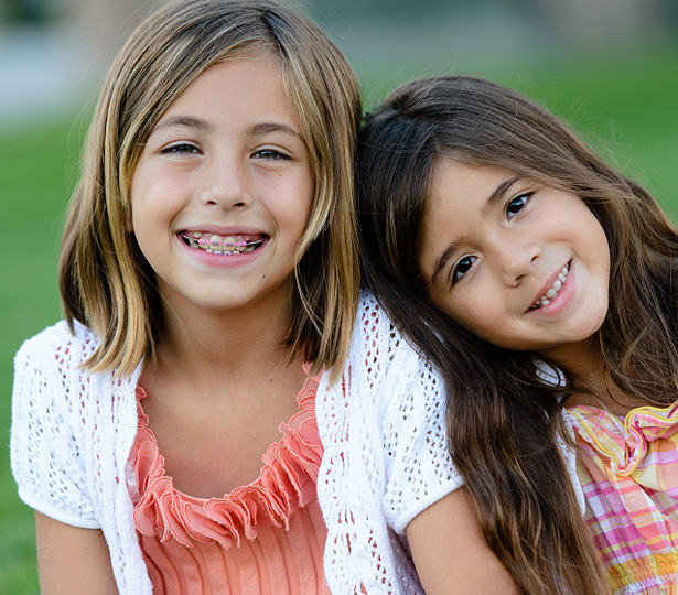 Two smiling girls posing together outdoors with green grass background.