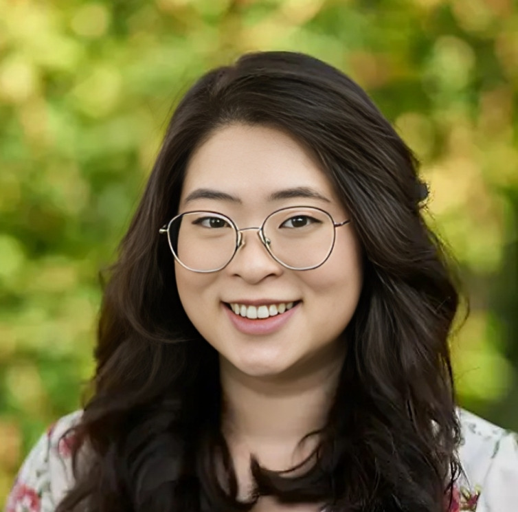 Young woman wearing glasses smiles at camera against blurred green background.