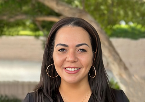 Professional headshot of smiling woman with dark hair and hoop earrings outdoors, dental office staff portrait.