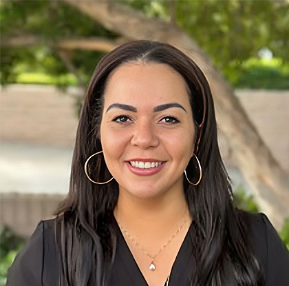 Professional headshot of smiling woman with long dark hair wearing hoop earrings and black shirt outdoors.