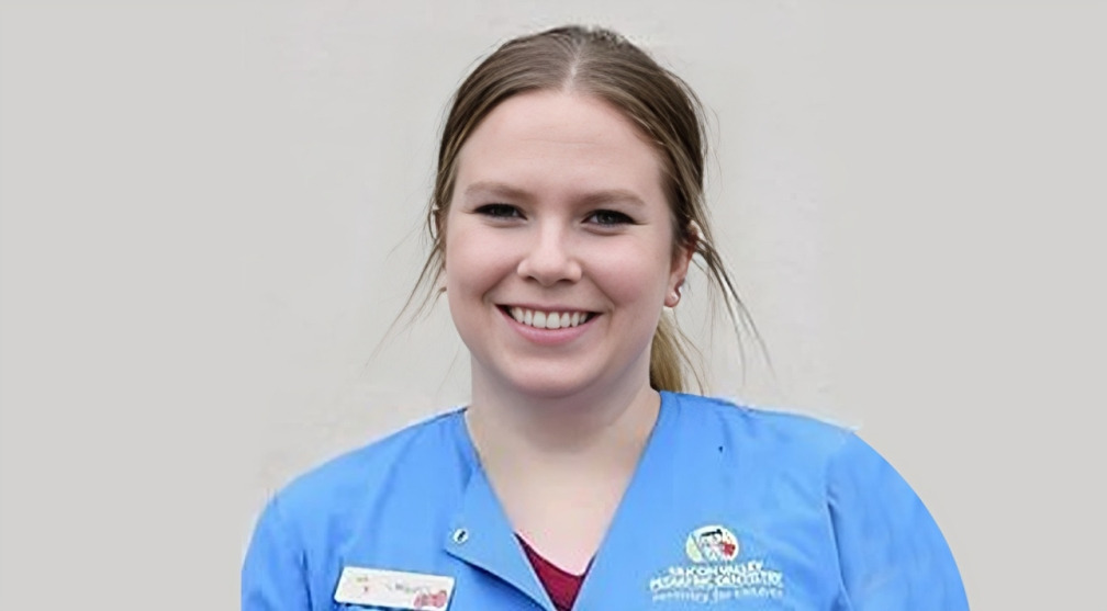 Smiling dental professional in blue scrubs wearing name badge against neutral background.