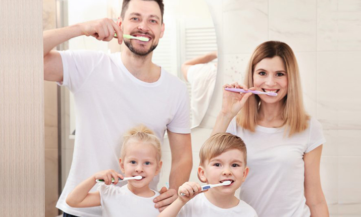 Happy family of four brushing teeth together in bathroom, smiling at camera.