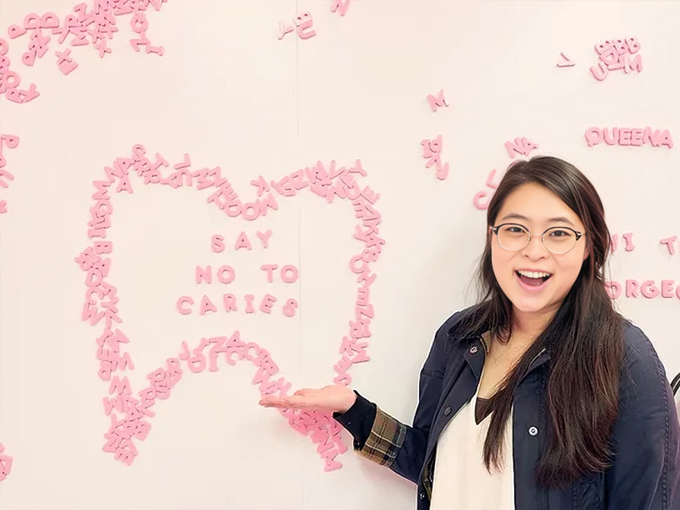Woman in dental office pointing to pink tooth design on wall with "Say No to Cavity" message.