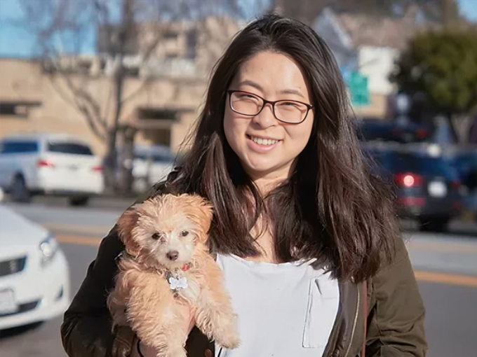 Woman wearing glasses holds a small brown dog outdoors in a parking lot setting.