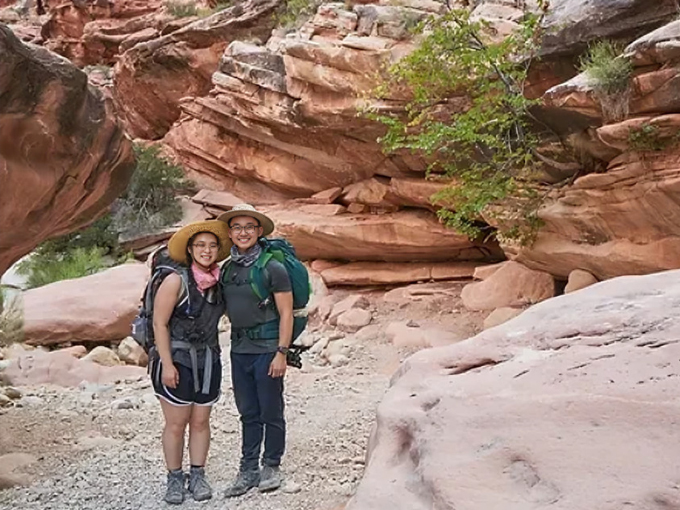 Two people hiking on red rock trail with desert landscape and vegetation.
