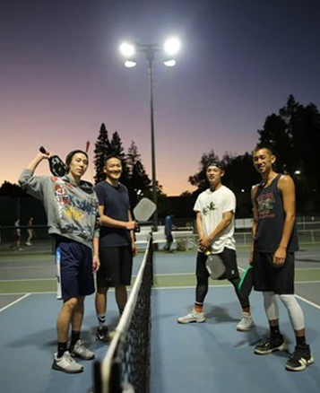 Five young people posing together on illuminated tennis court at dusk with net.