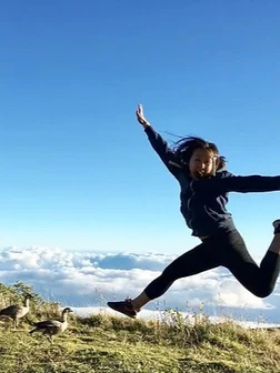 Child jumping joyfully outdoors on hillside with clear blue sky and clouds background.