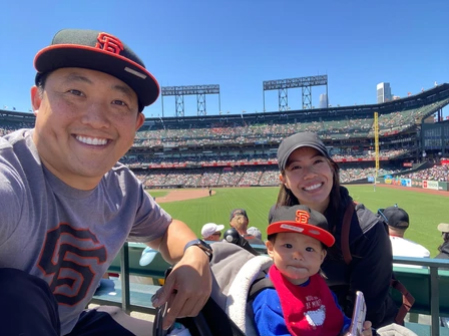 Family of four wearing red baseball caps at a baseball stadium during daytime game.