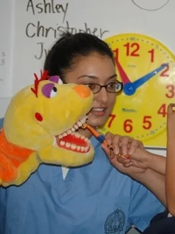 Young girl wearing glasses holds colorful toy chicken near yellow clock in dental office setting.