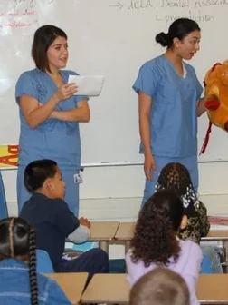 Two dental professionals in blue scrubs educate children in classroom setting with whiteboard.