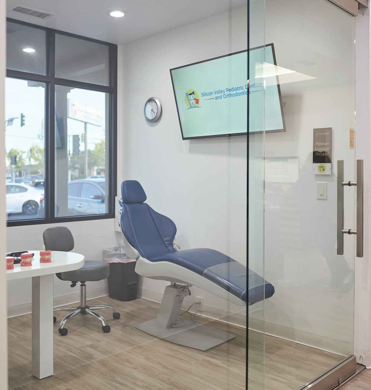 Modern dental operatory with blue patient chair, computer monitor, and glass partition in pediatric dental office.