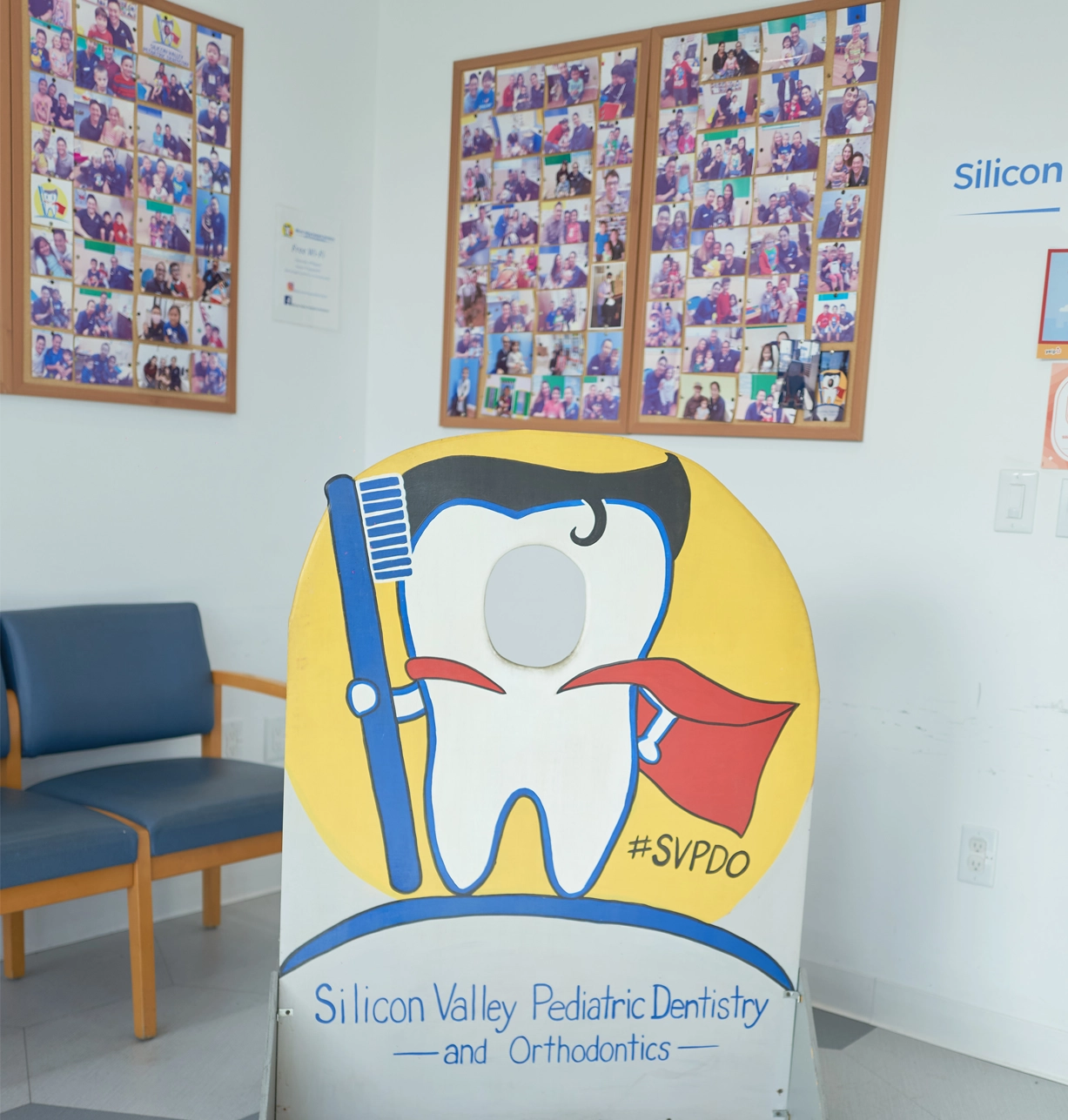 Cheerful tooth mascot holding toothbrush in pediatric dental office waiting room with photo boards.