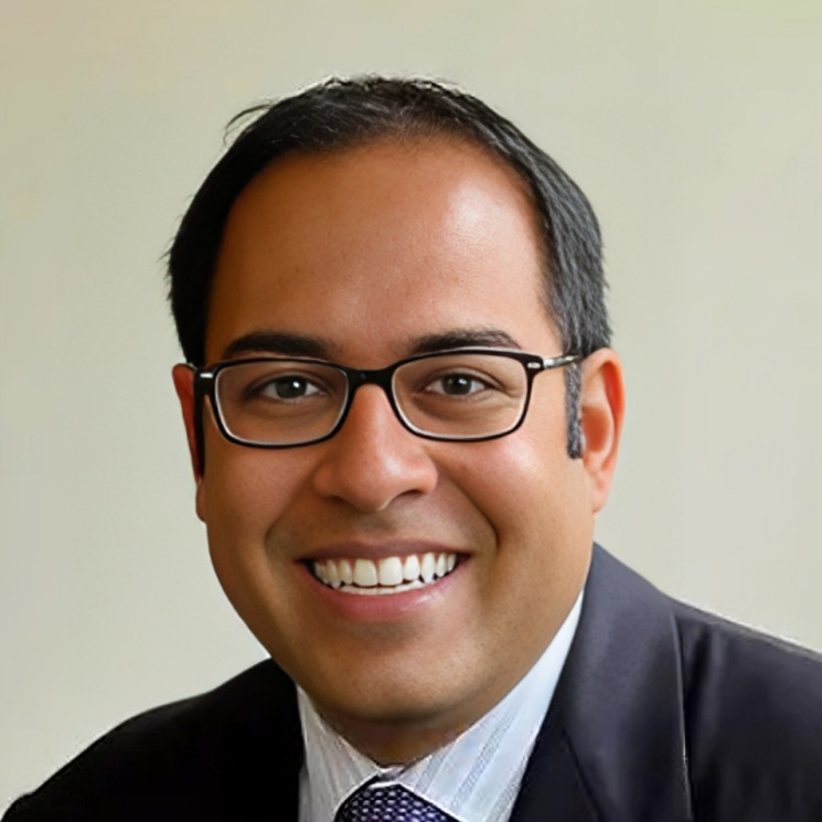 Professional headshot of male dentist wearing glasses and dark blazer in clinical office setting.