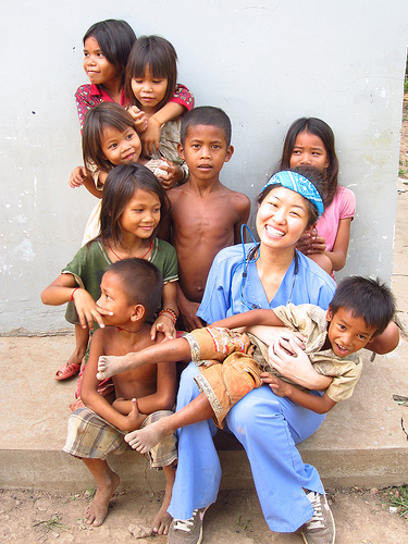 Healthcare worker in blue scrubs smiling with group of children in casual outdoor setting.