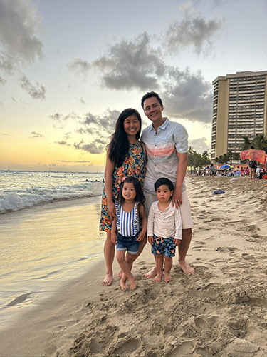 Family of four posing together on beach at sunset with high-rise building visible in background.