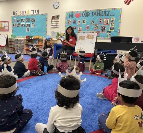 Teacher leads classroom activity with young children sitting on colorful mat wearing headbands.