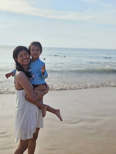 Woman holding young child on beach with ocean in background.