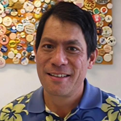 Male dentist smiling at camera wearing blue floral shirt, buttons backdrop visible.