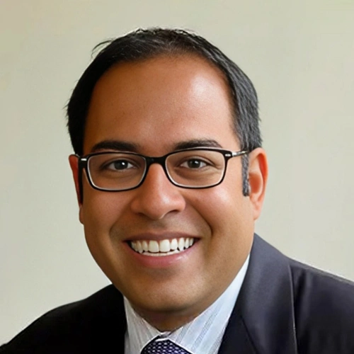 Professional headshot of a smiling male dentist wearing glasses and business attire against neutral background.