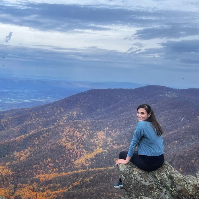 Woman sitting on mountain overlook wearing blue jacket, scenic forested valley landscape below