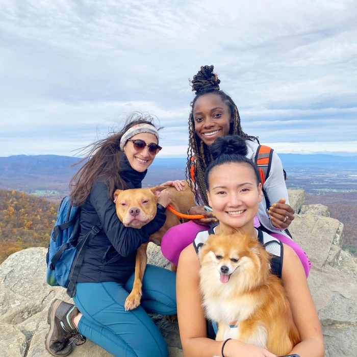 Three women and two dogs pose together on a mountain overlook with valley landscape in background.