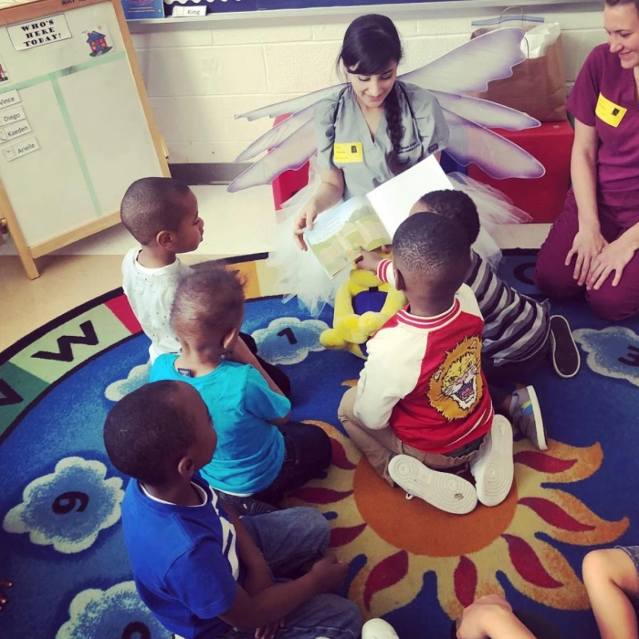 Young children sit in circle on colorful classroom floor mat during educational activity with adult instructors.