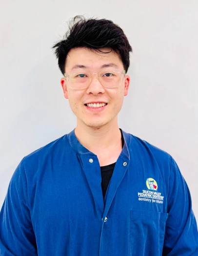 Professional headshot of smiling male dental staff member wearing blue clinic jacket against white background.