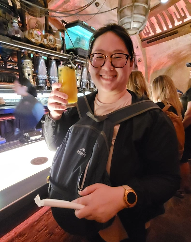 Woman holding beer at bar counter in casual indoor setting, smiling at camera.