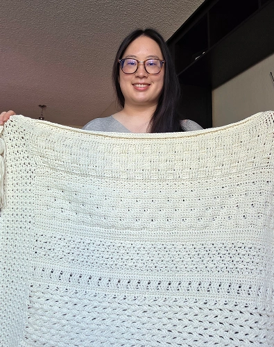 Woman holding white crocheted blanket indoors, smiling at camera wearing glasses.