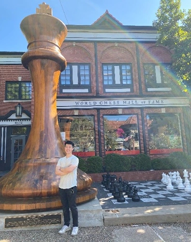 Woman posing next to giant wooden chess piece outside brick building with chess display.