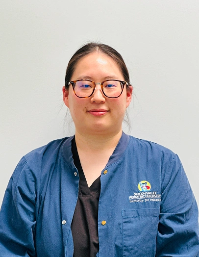 Female dental professional wearing blue scrubs and glasses, smiling at camera in clinical setting.