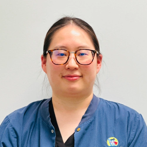 Woman wearing glasses and blue medical scrubs smiling at camera against white background.