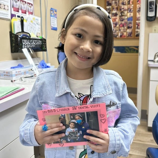 Young girl holding pink book in classroom, smiling at camera
