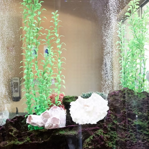 Decorative aquarium with white and pink fish ornaments among green aquatic plants and dark gravel.