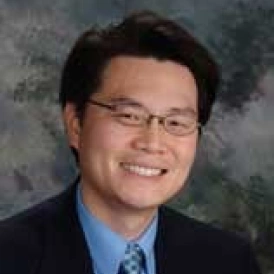 Professional headshot of an Asian male dentist wearing glasses and blue shirt against gray background.