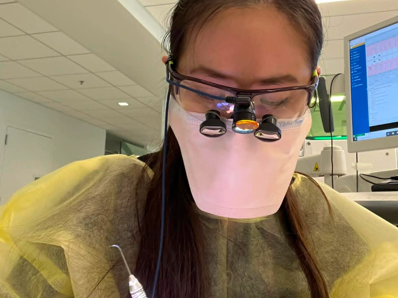A close-up clinical self-portrait of a dental professional wearing a medical mask, protective gown, and surgical loupes with an attached light.