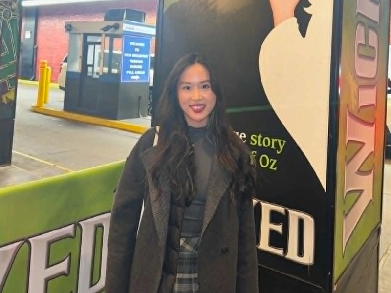 A young woman with long dark hair smiling in a grey coat in front of a green and black promotional display board for the musical Wicked.