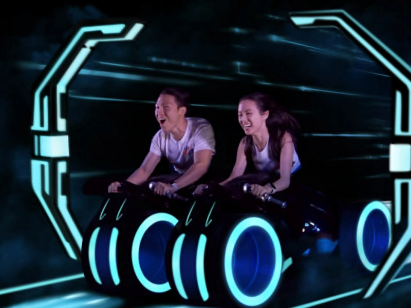 A young man and a young woman smiling and shouting with excitement while riding a TRON-style lightcycle roller coaster. They are leaning forward, gripping the handlebars of futuristic black motorcycles with glowing blue wheels, moving through a dark, high-speed tunnel with blue neon light streaks.
