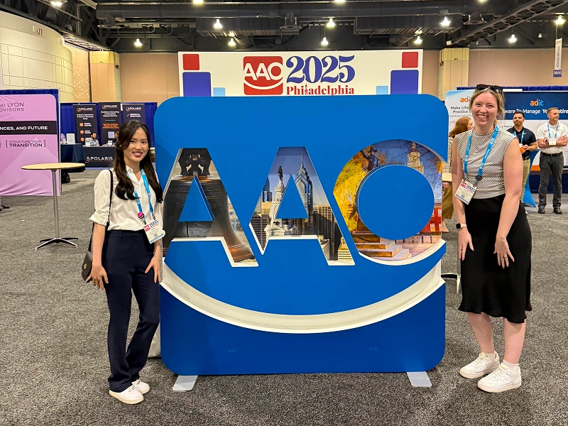 Two women smiling and posing on either side of a large blue and white "AAO 2025 Philadelphia" convention sign in an exhibition hall.