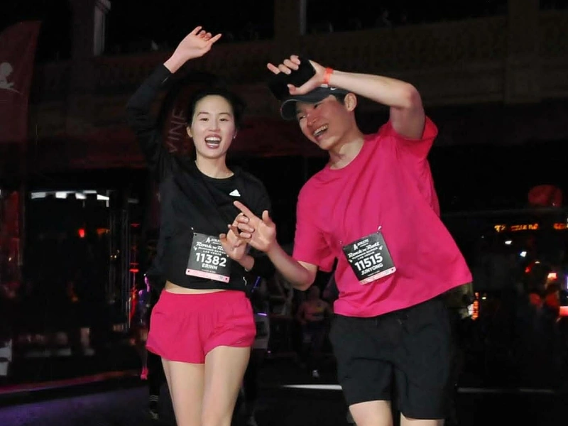 A young man and a young woman smiling and cheering mid-stride during a nighttime running event, wearing matching pink shorts and race bibs.