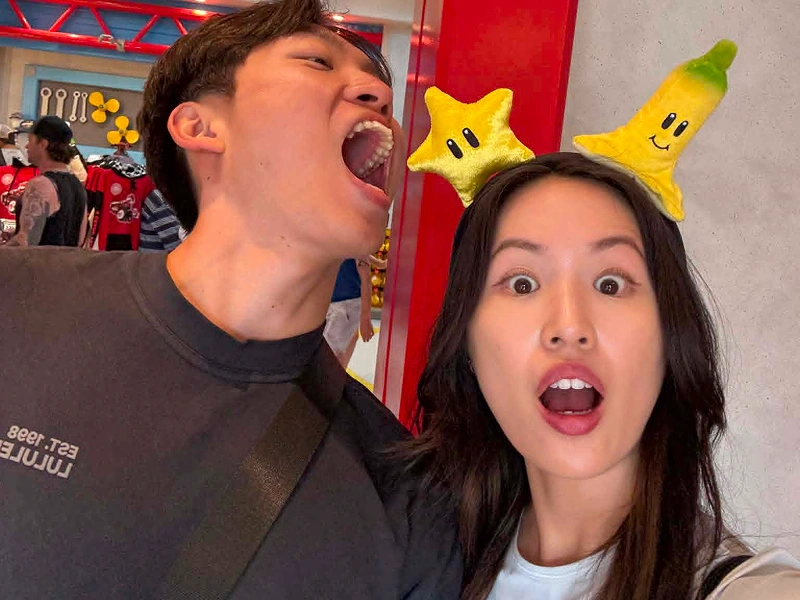 A close-up selfie of a young man with his mouth open in a playful shout alongside a young woman wearing bright yellow Nintendo star and banana headbands.