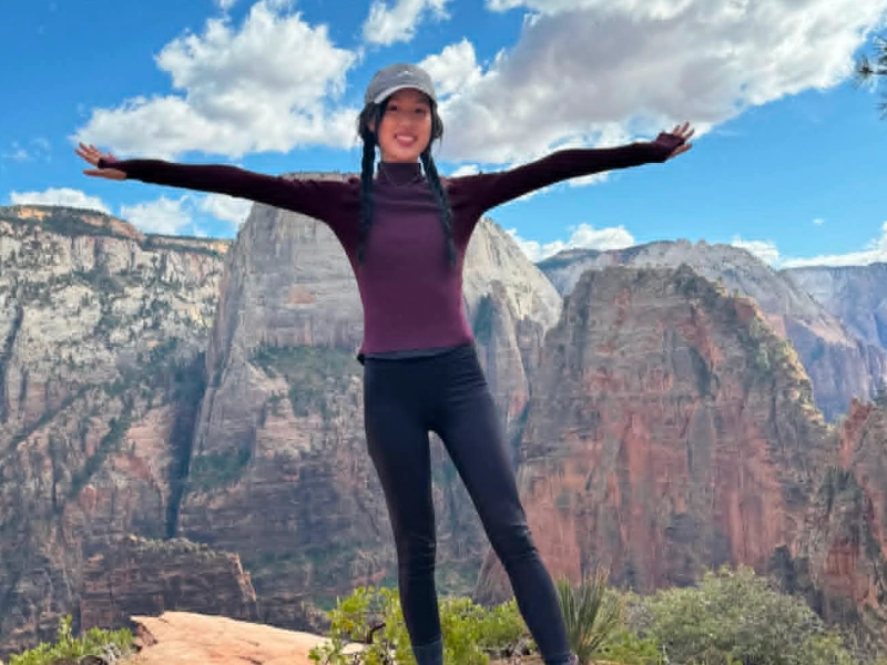 A woman standing at a mountain summit peak with arms wide open, wearing active-wear against a vast canyon and blue sky background.