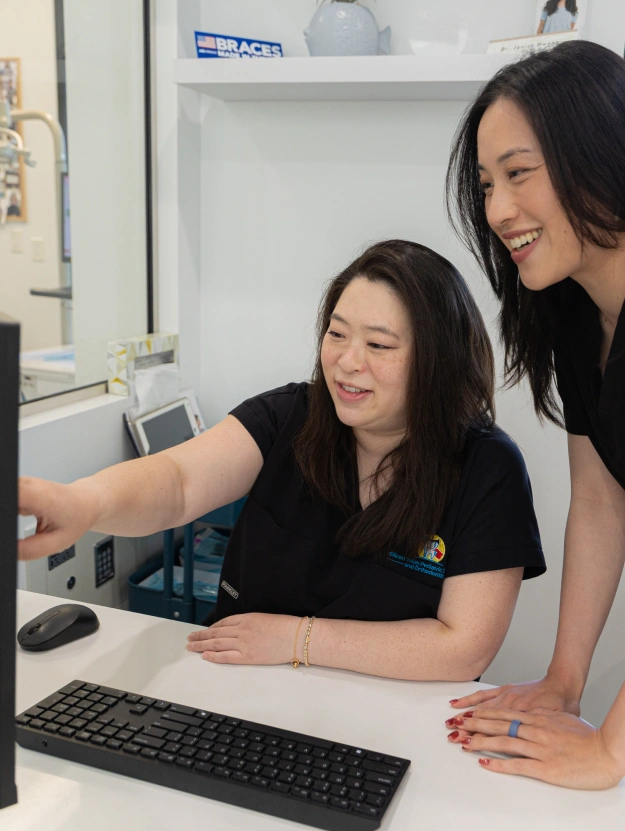 Two dental office staff members smile while reviewing patient information together on a desktop computer screen at a reception counter.