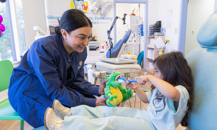 A pediatric dental provider sits next to a young patient in a dental chair, using a large toothbrush to demonstrate proper brushing technique on a green plush dinosaur toy.