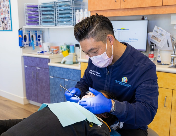 Dr. Scott Ngai, wearing a face mask and blue dental gloves, carefully performs a dental examination on a patient resting in a treatment chair.